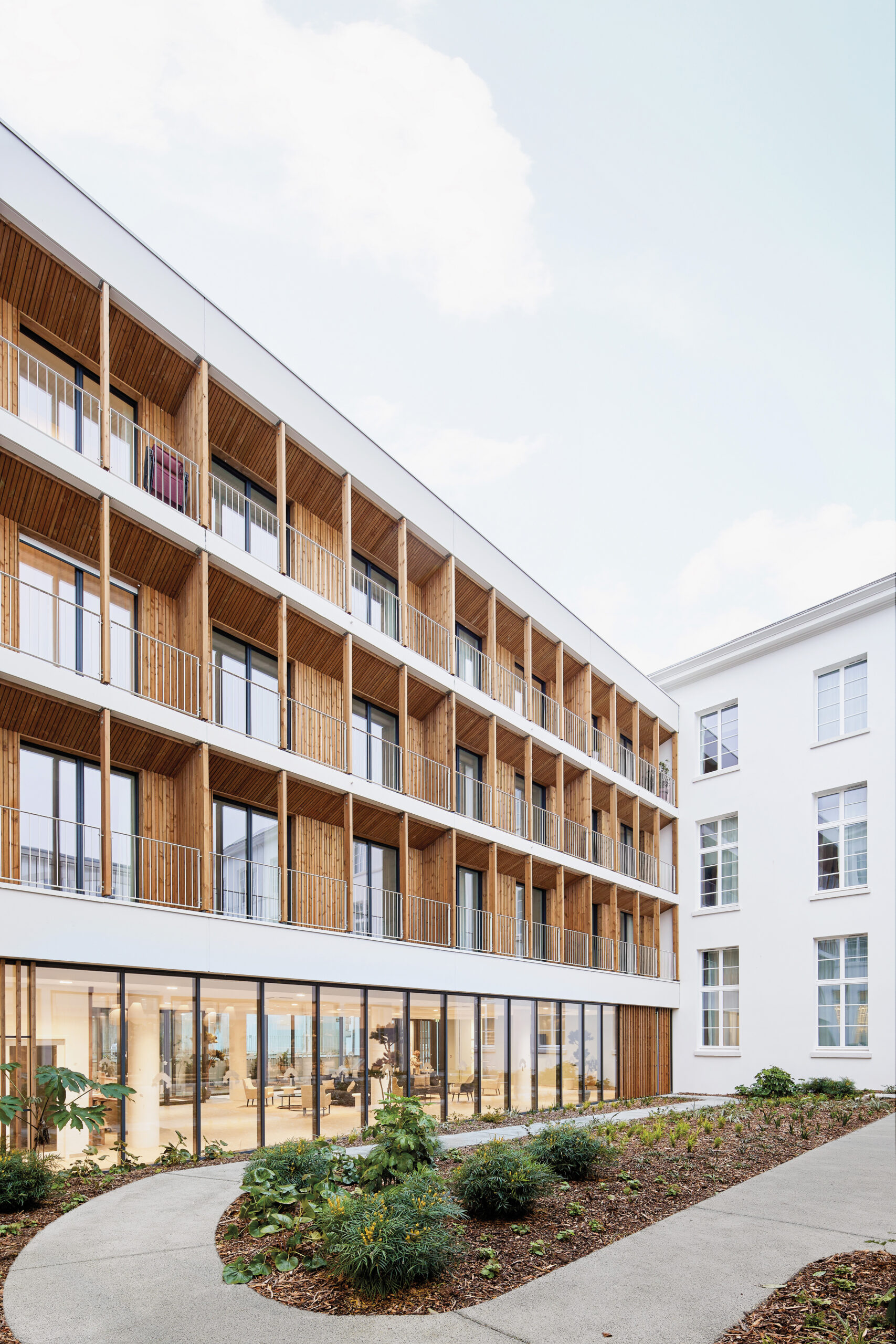 A woody exterior facade given to a senior living building with groud floor open to a courtyard full of greenery and covered in floor to ceiling glass. 