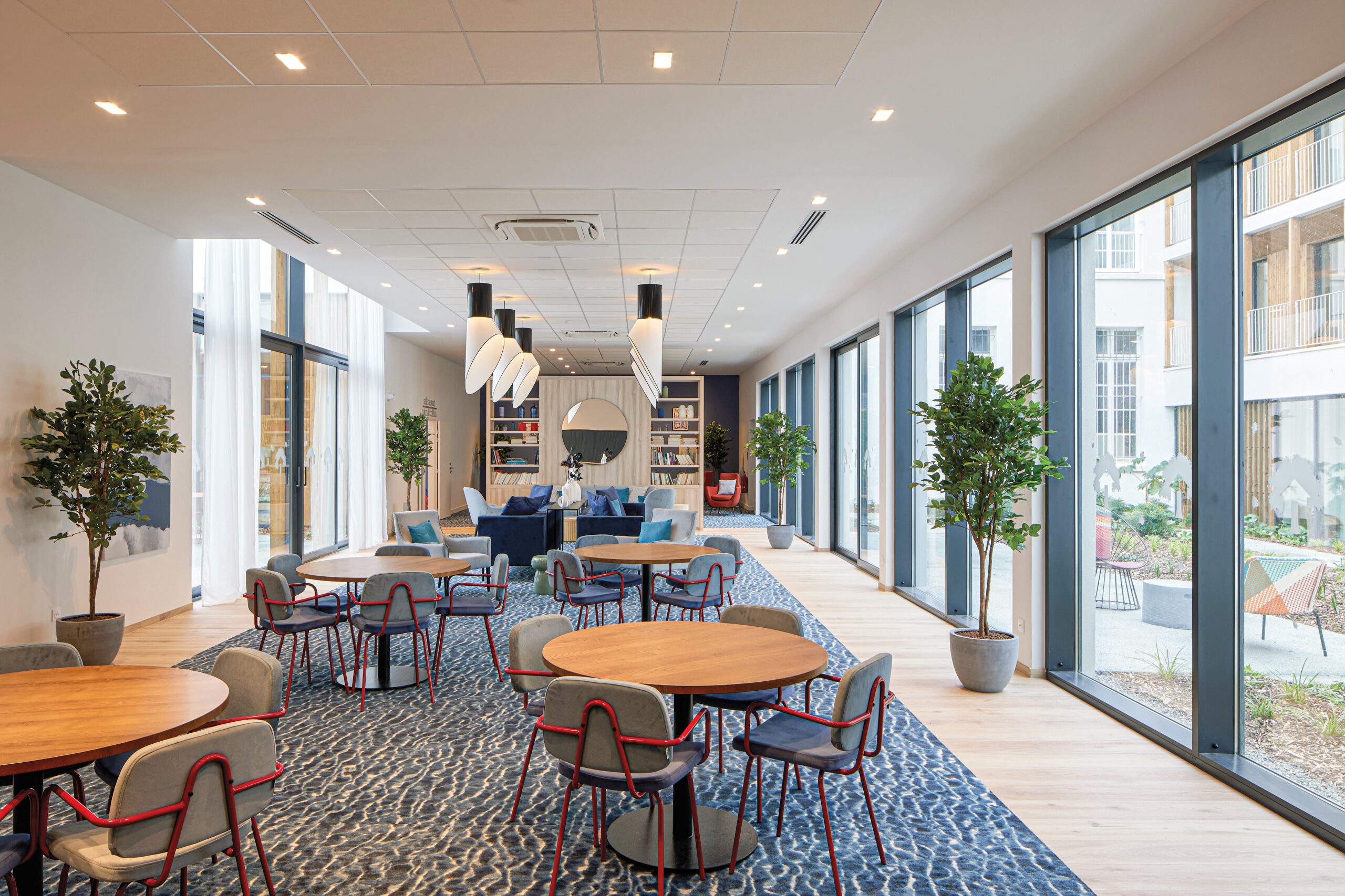 Interiors of the common lounge space with blue carpet, red and upholstered metal chairs and ergonomic seating with lots of natural light coming through the windows.