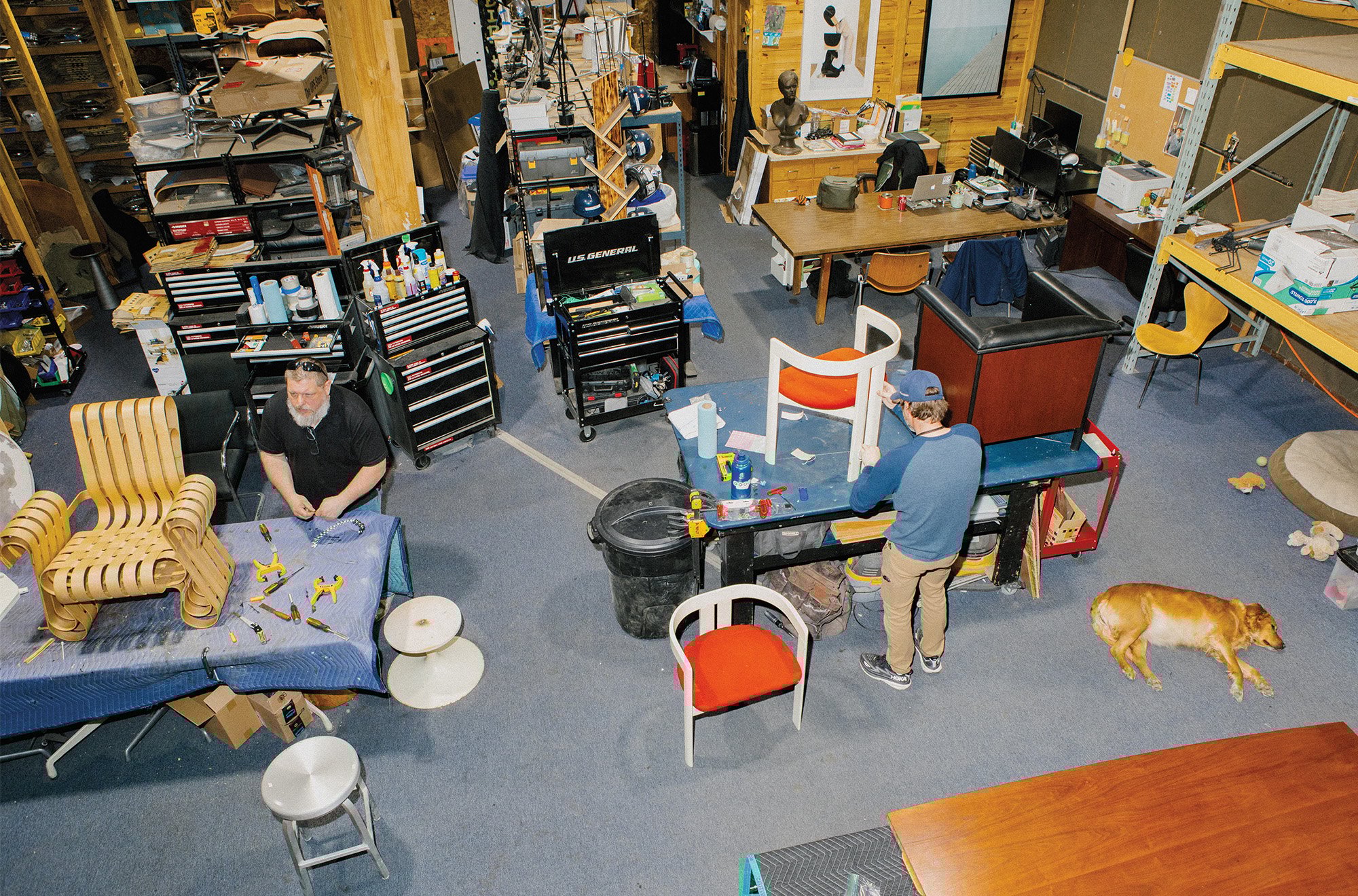 The shop floor where furniture makers are working on completing reclaimed and vintage pieces of furniture on their work tables and a dog lying on the corner. 