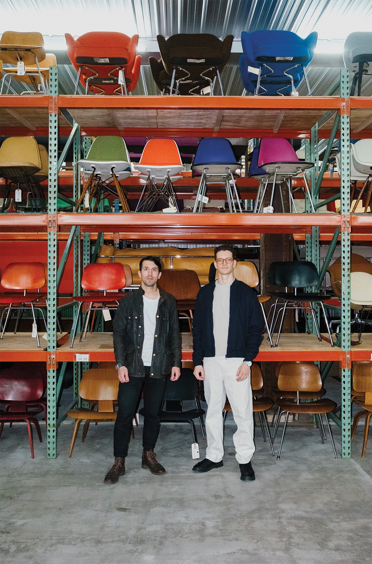 Cofounders standing in front of their acquired and collected chairs stacked and displayed on all shelves of the warehouse. 