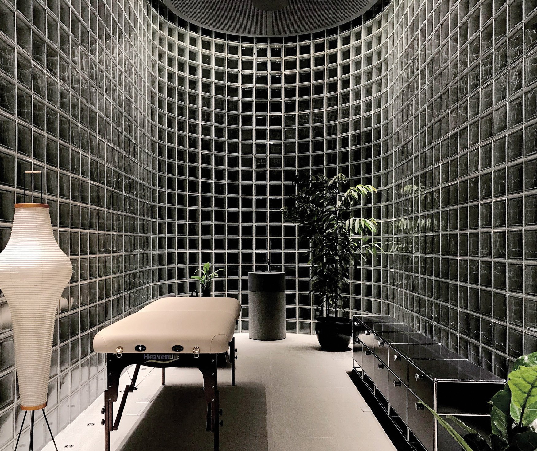 A curved space walled with glass blocks, a massage table, plants and a paper lamp kept inside. 