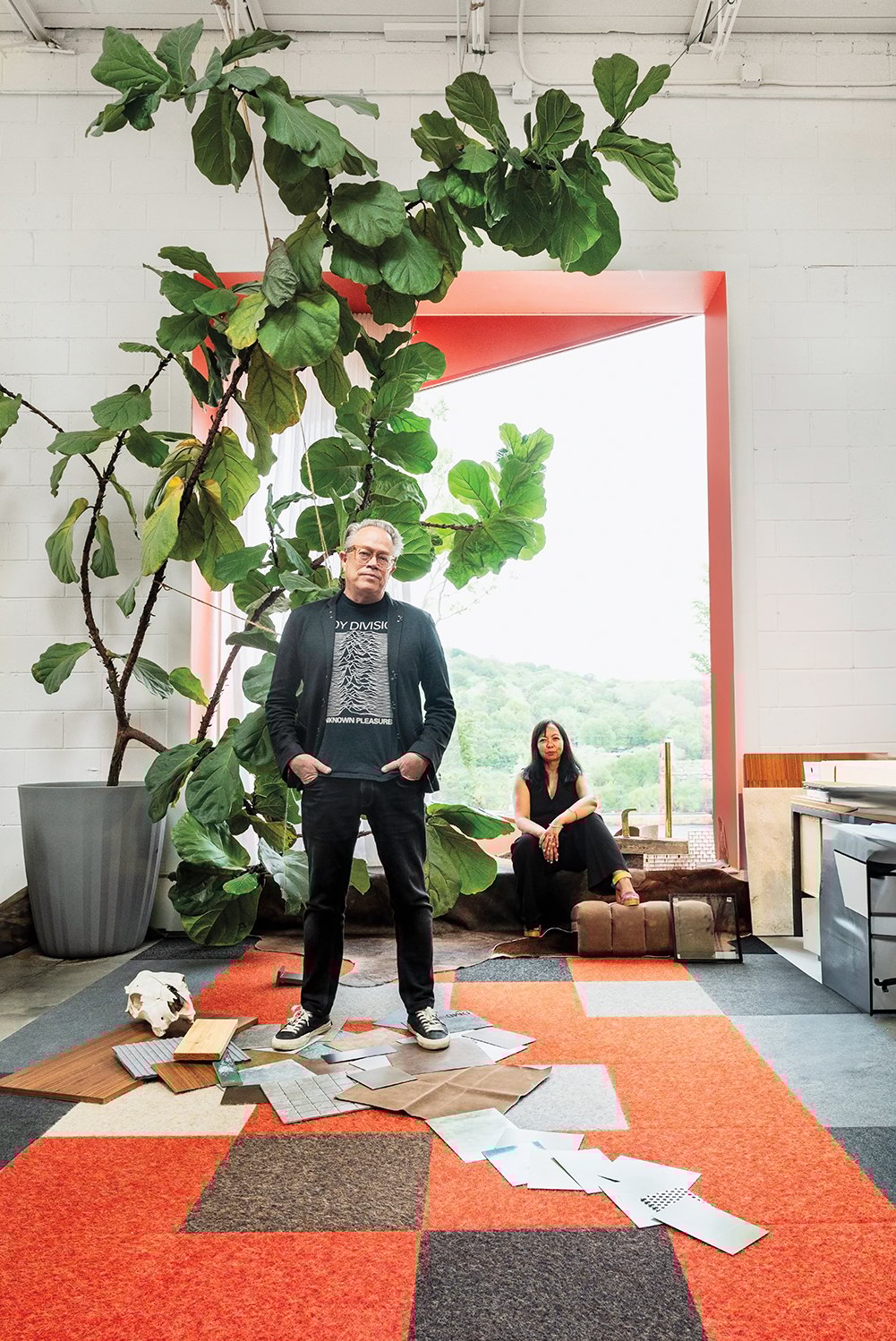 Marlon and Ati Blackwell pose in their studio with a large fiddle leaf fig plant and a red framed full length glass window. The orange checkered floor has a lot of scattered materials under his feet. 